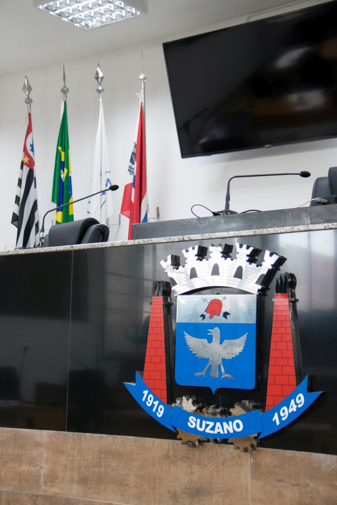 foto do brasão de Suzano em alto relevo, instalado em frente à Mesa Diretiva da Câmara de Suzano. Acima dele, estão hasteadas as bandeiras (da esq para a direita) do Estado de São Paulo, do Brasil, do Mercosul e de Suzano.