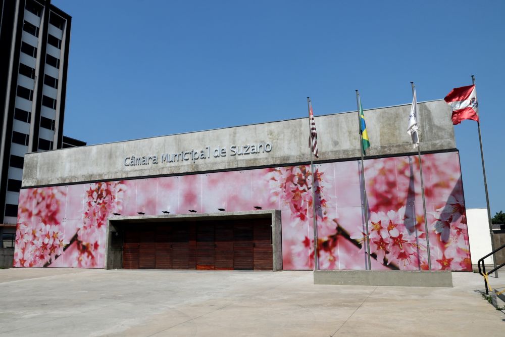 foto da entrada do Plenário da Câmara de Suzano, a fachada é adesivada com imagens de flores de cerejeiras, na imagem aparecem as bandeiras hasteadas do Brasil, São Paulo, Mercosul e Suzano, na foto o céu está bastante azul
