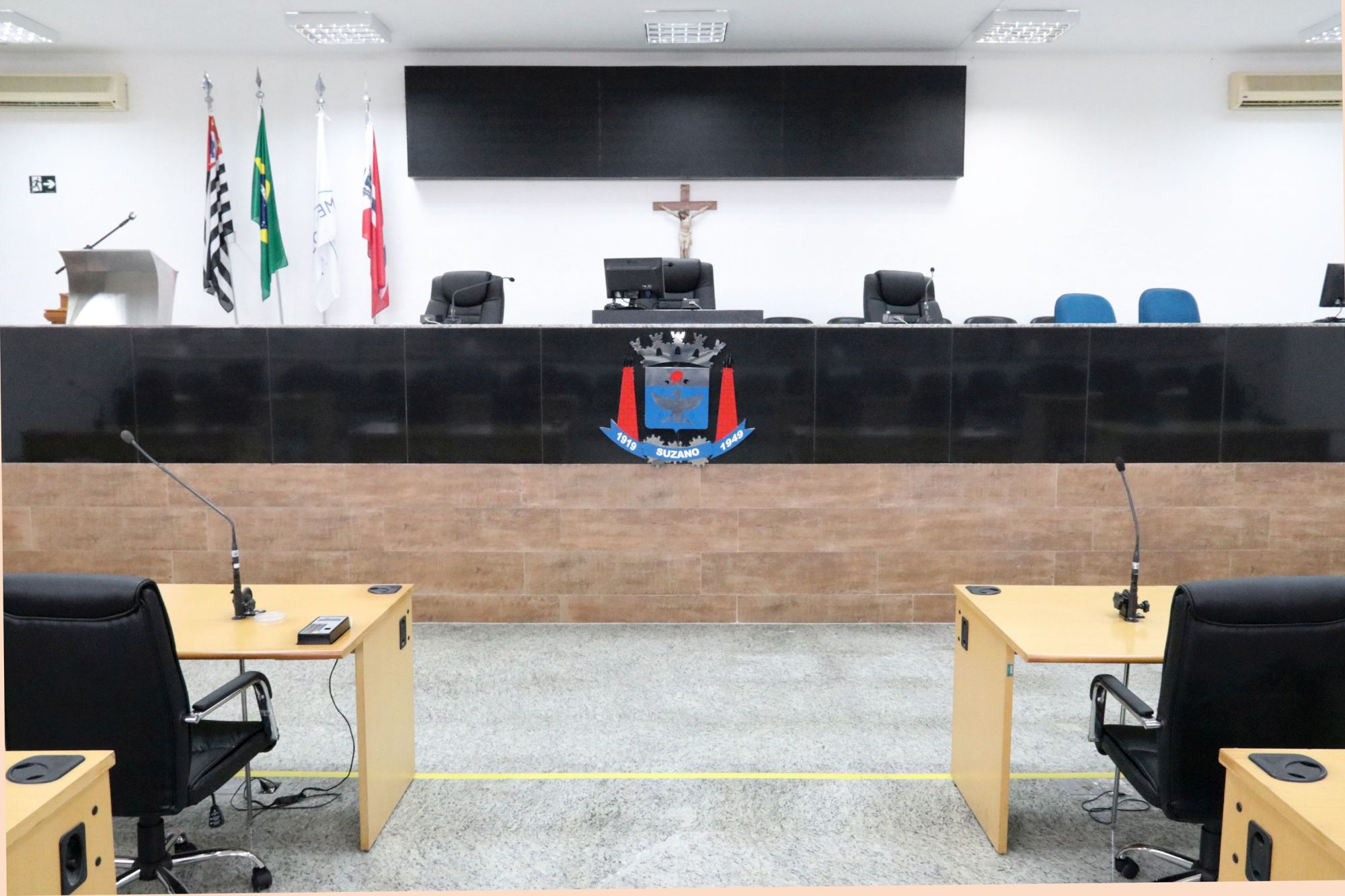 foto da mesa diretiva no Plenário da Câmara de Suzano, ao fundo painel de votação, as bandeiras do Brasil, São Paulo e Suzano, e a tribuna onde são feitos os discursos dos vereadores durantes as sessões