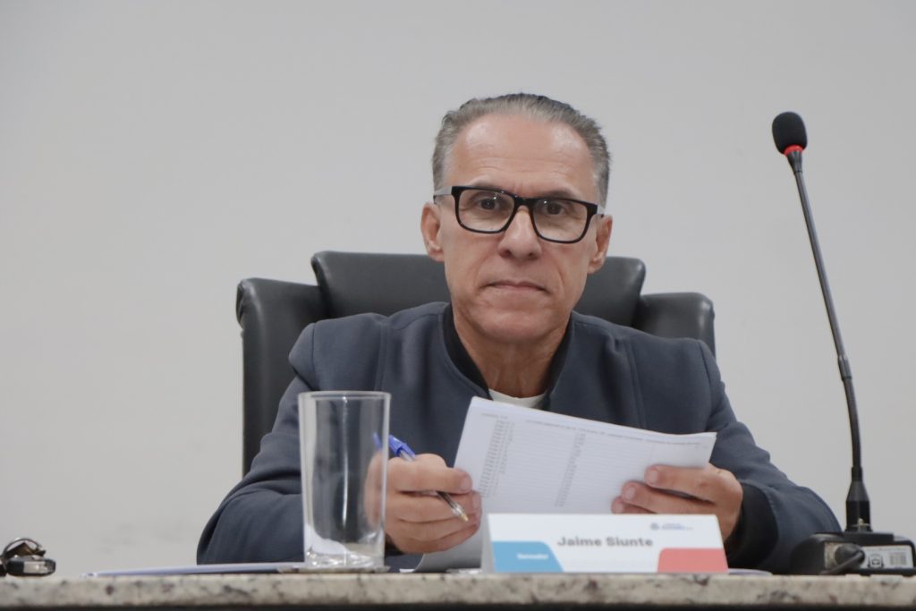 vereador Jaime Siunte sentado na Mesa Diretiva da Câmara segura um papel com as mãos. Ele usa óculos e uma jaqueta cinza.