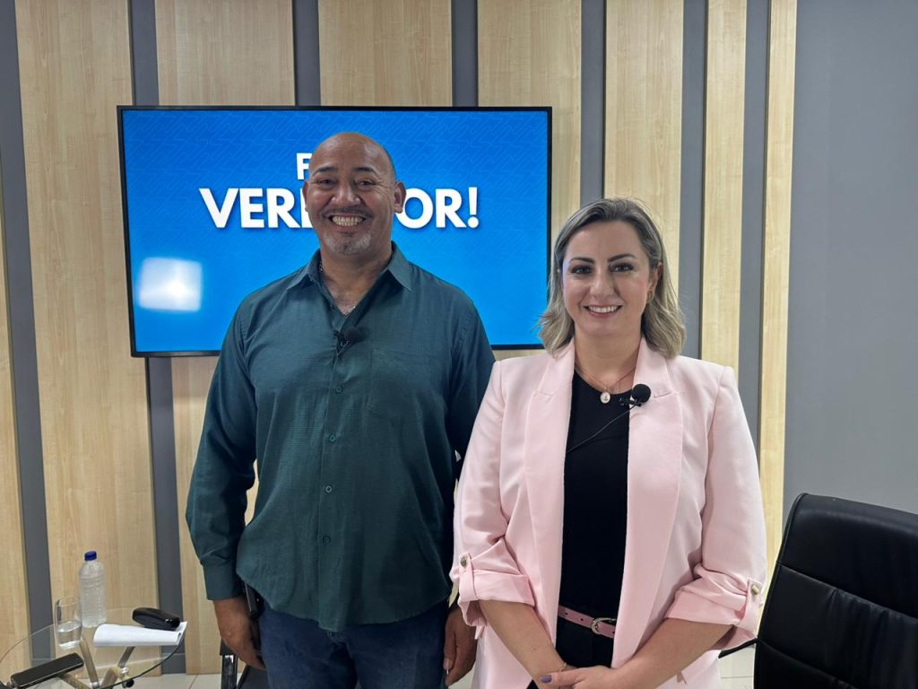 ereador João Sabugo e ex-primeira-dama Larissa Ashiuchi lado a lado no estúdio da TV Câmara Suzano. Ambos sorriem para a foto.