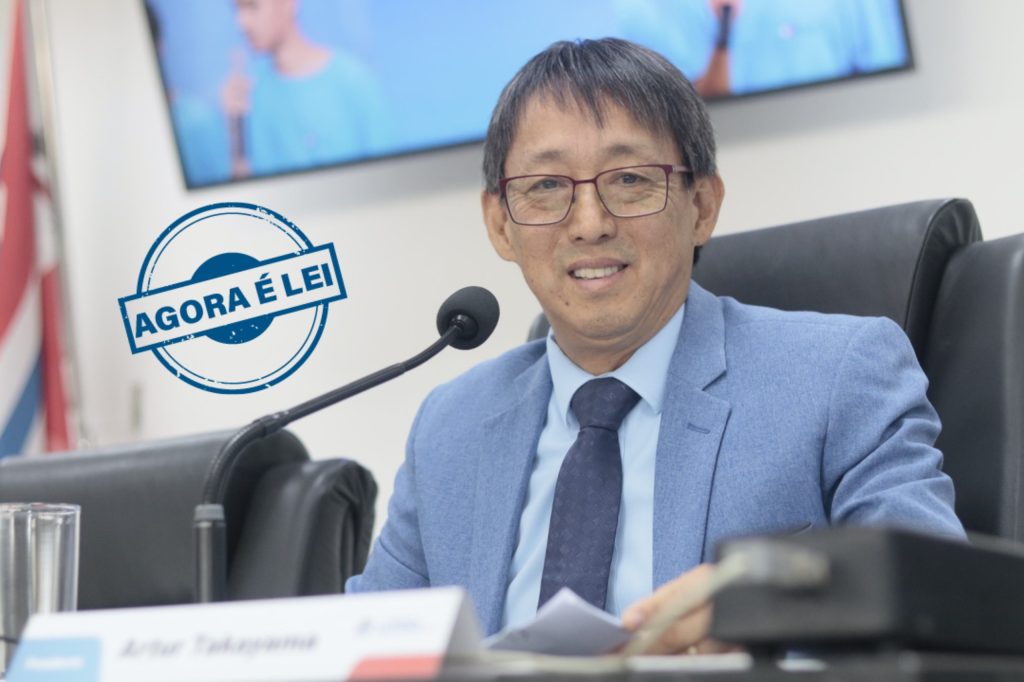 Vereador Artur Takayama sentado na cadeira da Presidência da Câmara. Ele sorri para a foto e usa terno e gravata azuis.