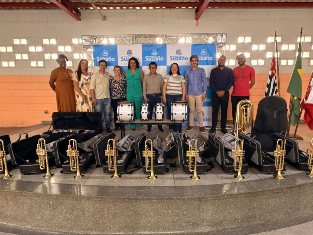 Autoridades municipais, educadores e representantes do projeto posam para foto durante a entrega de novos instrumentos musicais a uma escola da rede municipal de Suzano. Em primeiro plano, estão dispostos diversos instrumentos de sopro e percussão, que serão utilizados nas atividades de musicalização dos alunos. Ao fundo, o grupo está reunido diante de um painel institucional da Prefeitura de Suzano.