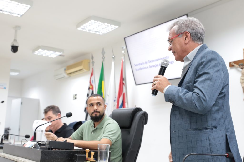 De pé, em primeiro plano, secretário Mauro Vaz fala ao microfone. Ele é observado pelo vereador Dirceu, que está sentado na Mesa Diretiva ao lado do diretor Arrones Júnior.