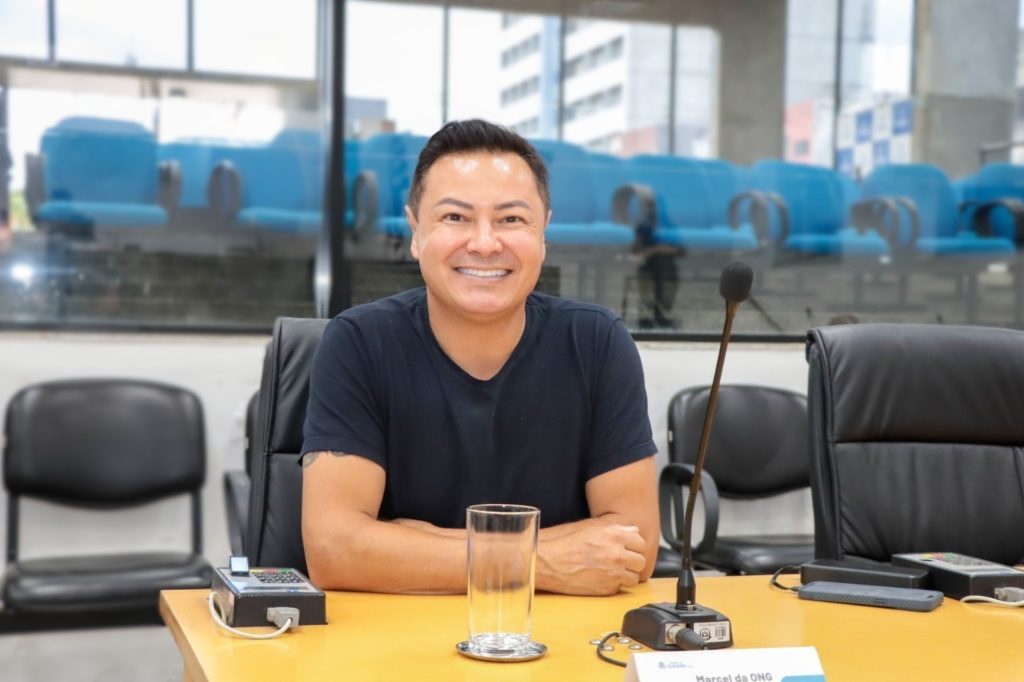 Vereador Marcel da ONG sentado em sua mesa no Plenário. Ele usa uma camiseta escura e sorri para a foto.