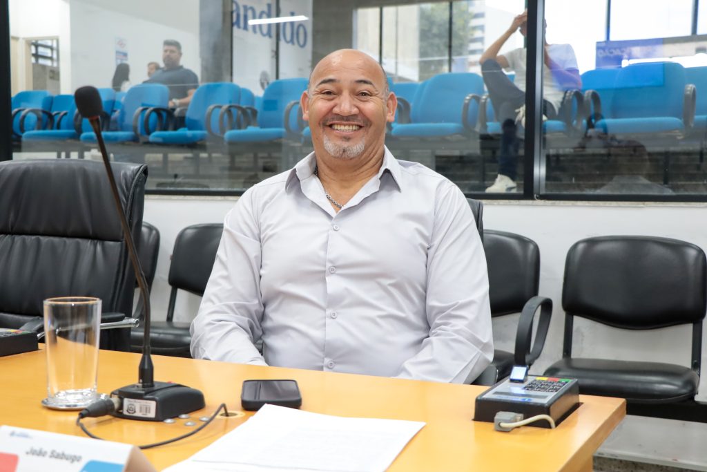 Sentado atrás de sua mesa no Plenário, vereador João Sabugo sorri para a foto. Ele usa camisa social clara.