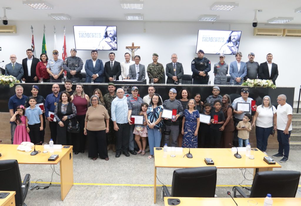De pé, no Plenário da Câmara, homenageados mostram suas medalhas ao lado de familiares. Acima deles, na Mesa Diretiva, estão vereadores, prefeito e demais autoridades presentes ao evento.