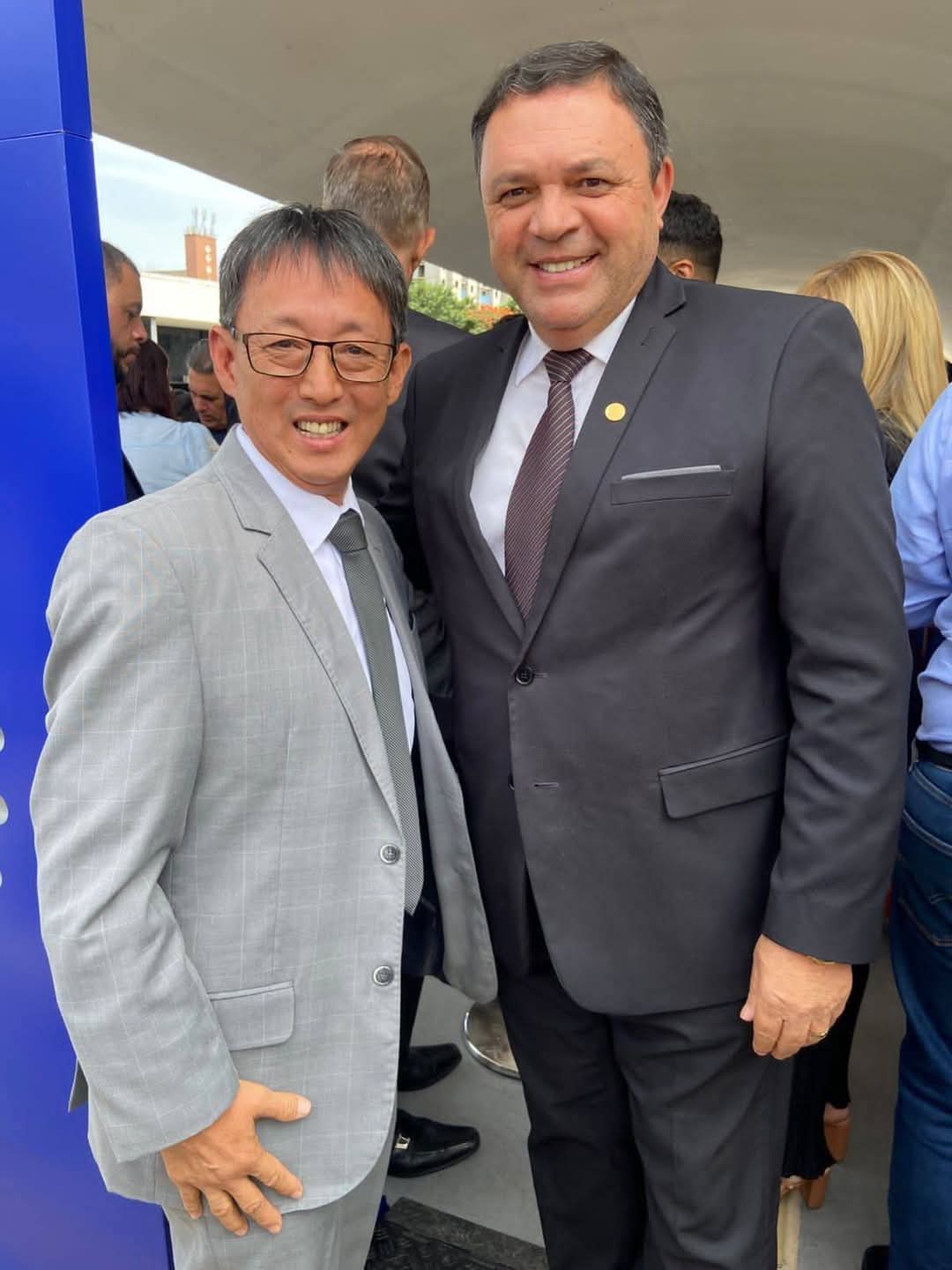 De pé, lado a lado, estão, da esquerda para a direita, Artur Takayama, de terno cinza e óculos, e Marcos Damásio, de terno preto.