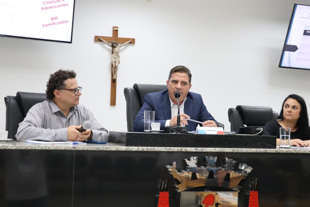 Sentados na Mesa Diretiva do Plenário, estão, da esquerda para a direita, o secretário Geraldo Garippo, o vereador Leandrinho e a diretora de Proteção Especial da Prefeitura Regiane Borges.