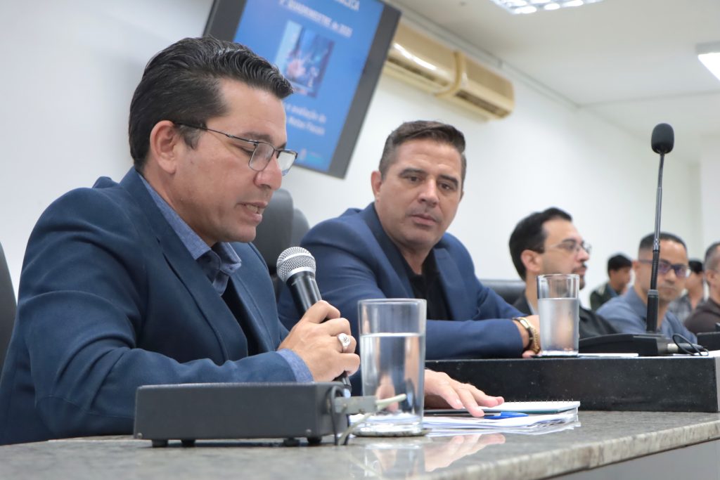 Em primeiro plano, secretário de Finanças, Itamar Viana, fala ao microfone, enquanto é observado pelo vereador Leandrinho. Ambos estão na Mesa Diretiva da Câmara em audiência realizada em 26 de fevereiro.