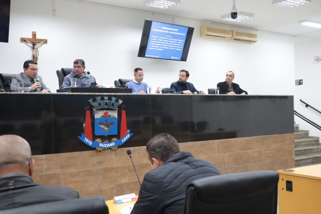 Em primeiro plano, de costas, vereadores Josias Mineiro e Leandrinho sentados no Plenário. Em segundo plano, de frente para a câmera, estão na Mesa Diretiva, da esquerda para a direita, o secretário Itamar Viana, o vereador Denis e os contadores Claudinei Estevão, Marcelo Rodrigues e Maurício Bento.
