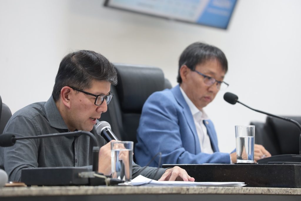 sentados na Mesa Diretiva da Câmara, o secretário William Harada fala ao microfone, tendo ao seu lado o vereador Artur Takayama.