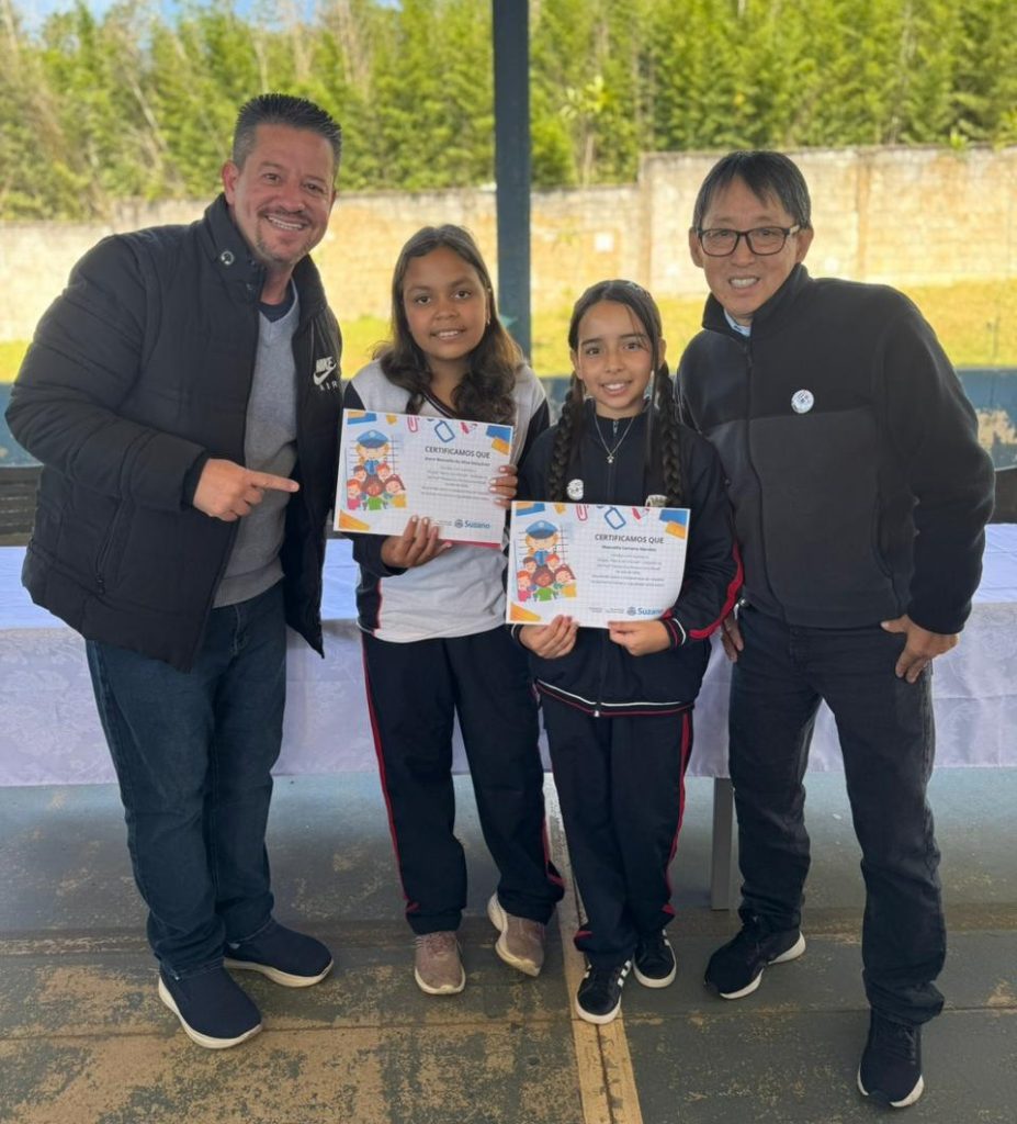 Vereador Maizena (à esquerda da foto) e vereador Artur Takayama (à direita) ladeiam duas meninas que seguram certificados.