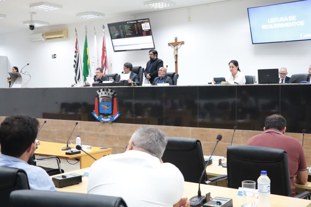 Em primeiro plano, de costas, vereadores sentados em suas mesas no Plenário. À frente deles, estão os vereadores da Mesa Diretiva.