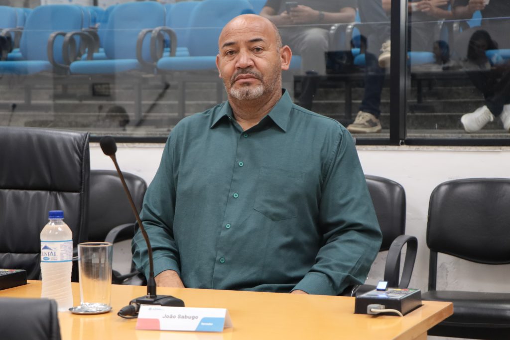 vereador João Sabugo sentado em sua mesa no Plenário. Ele usa uma camisa social verde.