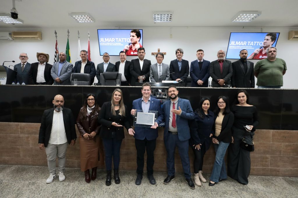 De pé, no Plenário da Câmara de Suzano, vereador Jilmar Tatto segura a placa com o Título de Cidadão Suzanense ao lado do vereador André Dourado e outras pessoas presentes ao evento. Acima deles, na Mesa Diretiva, estão o prefeito, vereadores e demais autoridades presentes à sessão.
