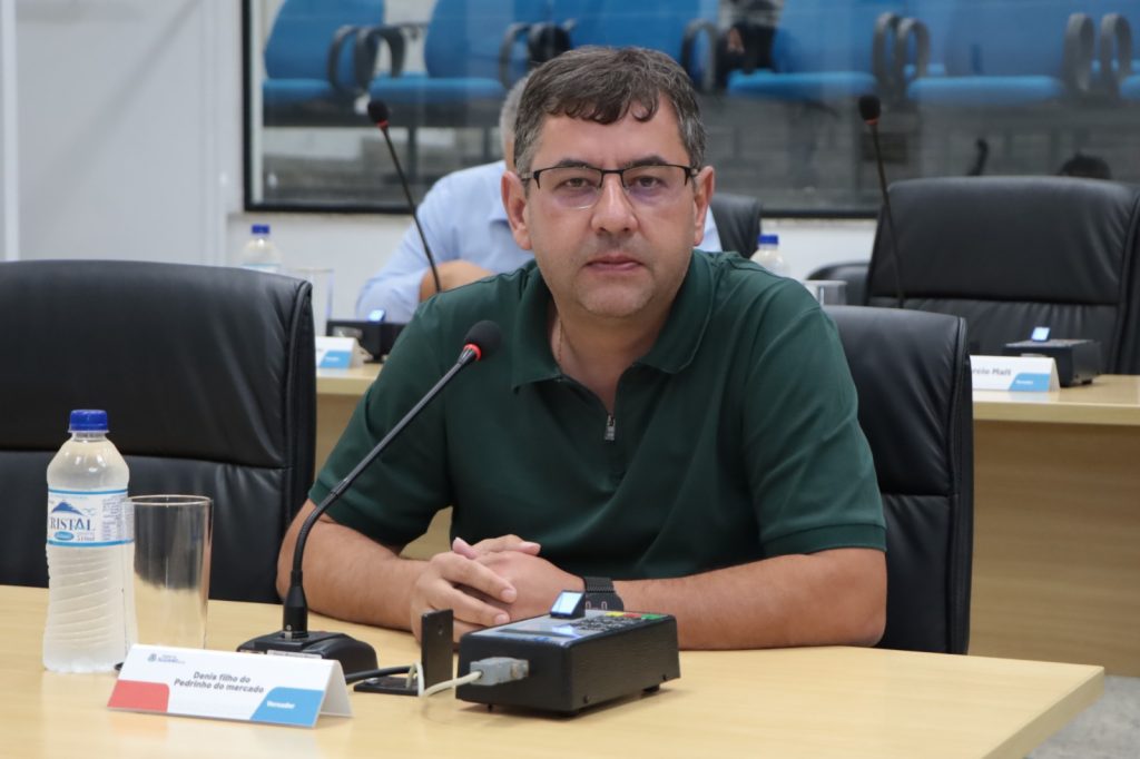 Vereador Denis sentado atrás de sua mesa no Plenário. Ele usa óculos e uma camisa polo verde.
