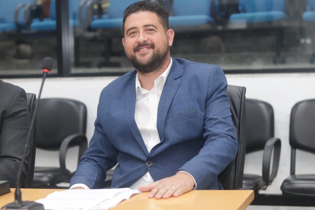 Vereador André Dourado sentado atrás de sua mesa no Plenário da Câmara. Ele usa blazer azul sobre camisa social branca e sorri para a foto.