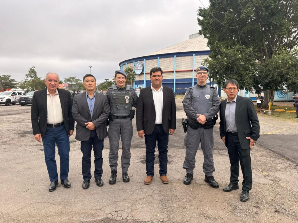 De pé, tendo ao fundo a Arena do Parque Max Feffer, estão o vereador Artur Takayama (primeiro à direita) e o prefeito Pedro Ishi (terceiro à direita) entre policiais e civis presentes na foto.