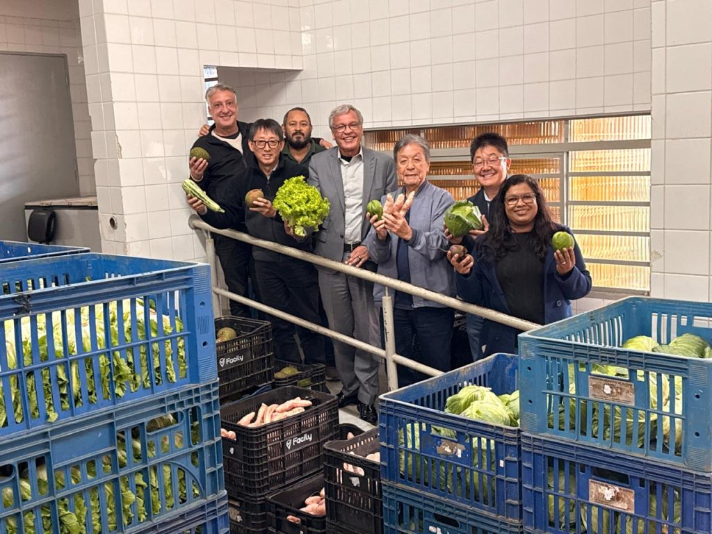 vereador Artur Takayama (segundo à esquerda, de óculos), secretário Mauro Vaz (ao centro, de blazer cinza e óculos) posam para foto segurando verduras e legumes junto a outros funcionários do local. À frente deles, estão várias caixas plásticas empilhadas com verduras e legumes.