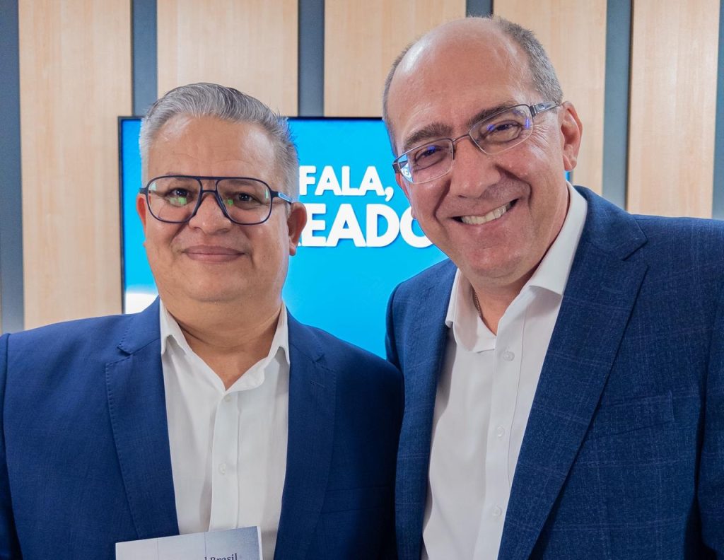 No estúdio da TV Câmara Suzano, vereador professor Edirlei (à esquerda) e professor Leandro Bassini posam para foto. Ambos usam óculos, camisa social branca e blazer azul.