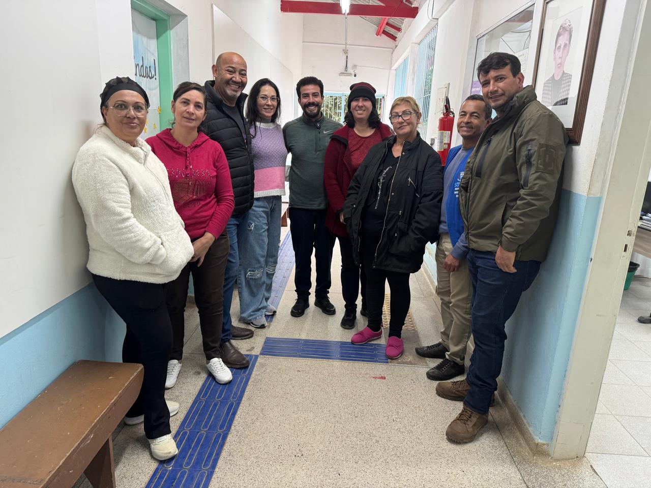 de pé, em um corredor da escola, estão lado a lado, em formato de meia-lua, funcionários da escola e os vereadores João Sabugo (terceiro à esquerda), Marcio Malt (ao centro) e Adilson Horse (primeiro à direita).