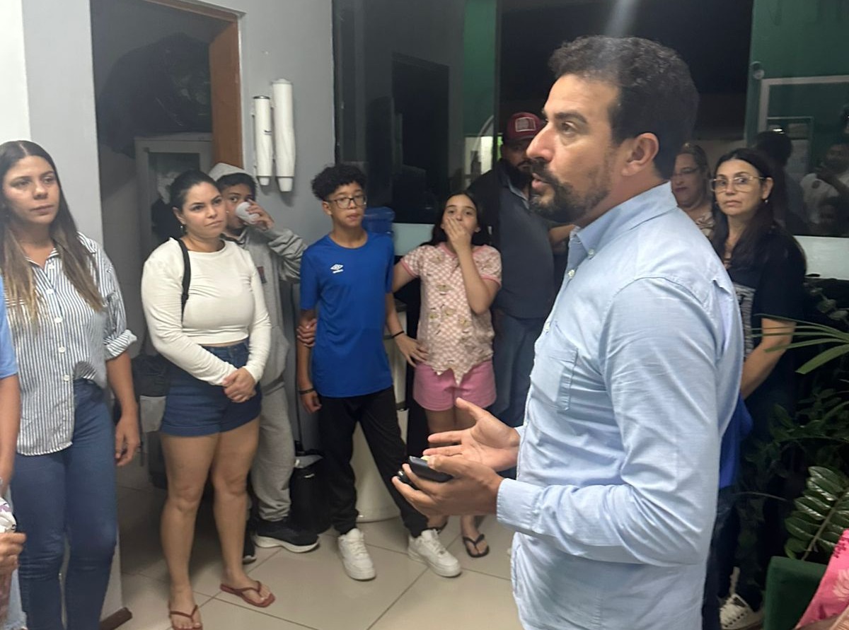 vereador Marcio Malt fala em pé para um grupo de moradores reunidos em um ambiente interno. Ele gesticula com as mãos enquanto as pessoas ao redor observam atentamente. Entre os presentes há homens, mulheres e adolescentes, alguns em pé próximos à porta e às paredes do local.