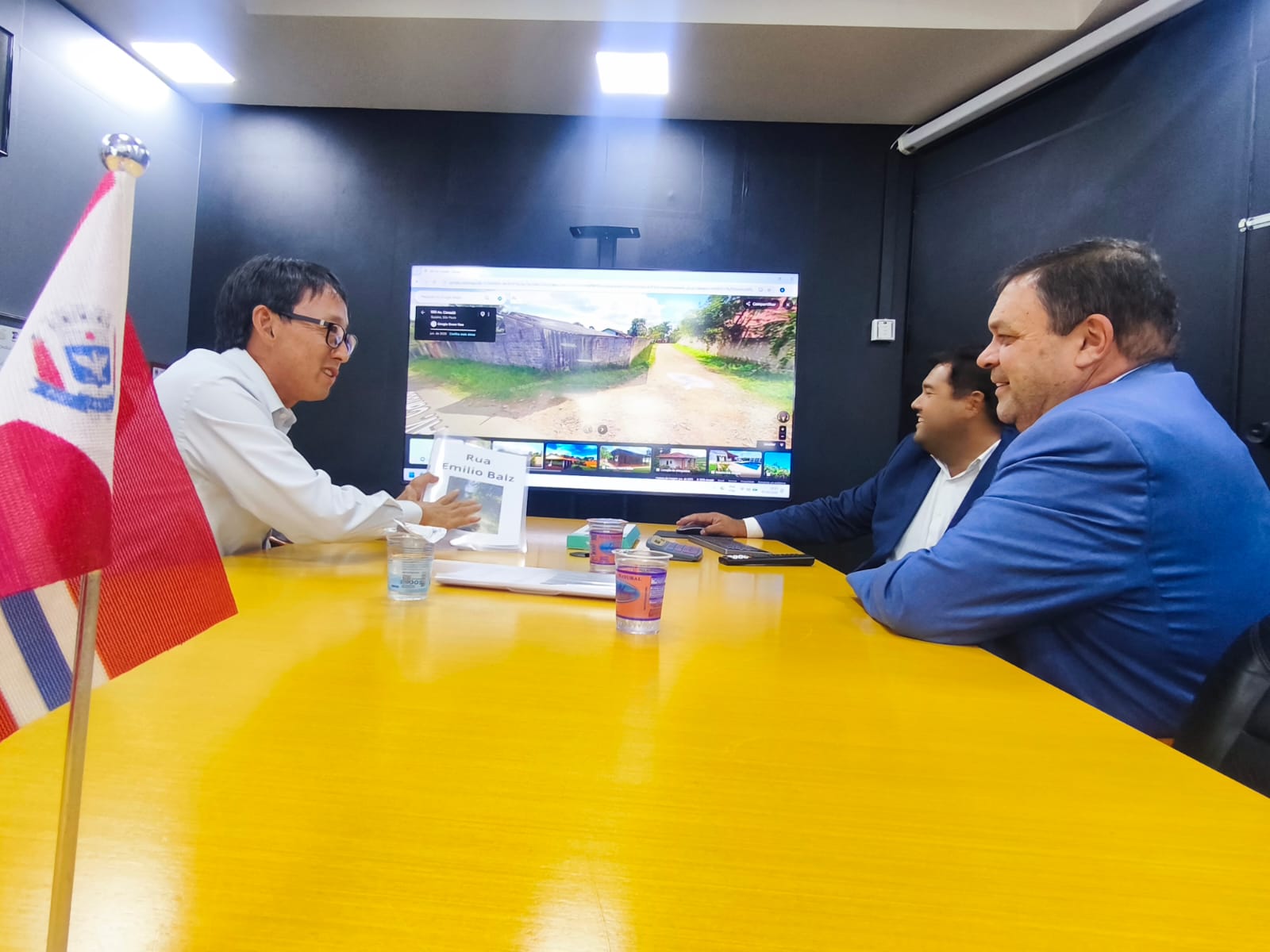   vereador Artur Takayama, prefeito Pedro Ishi e deputado estadual Marcos Damasio participam de reunião em uma sala com mesa amarela. Ao fundo, uma televisão exibe a imagem de uma rua em mapa digital.