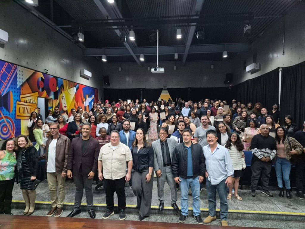 no anfiteatro Wilma Bentivegna, estão formandas de pé ao fundo e autoridades à frente. Ao centro, está o presidente da Câmara, Artur Takayama, de óculos e terno e gravata.