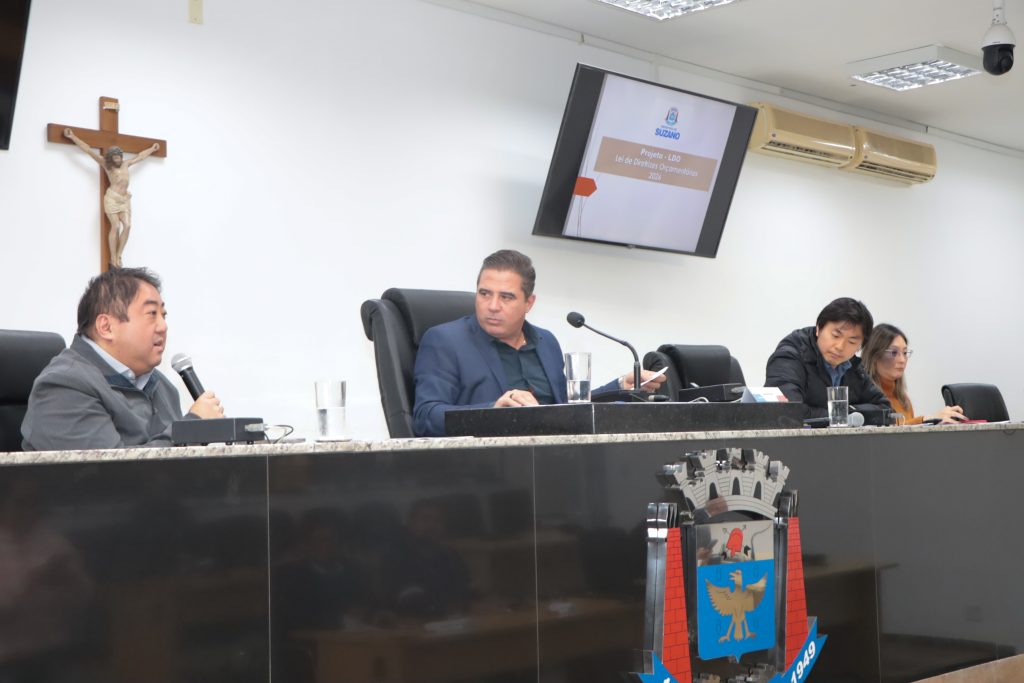 na Mesa Diretiva da Câmara, o secretário de Planejamento Denis Watanabe fala ao microfone enquanto é observado pelo vereador Leandrinho em audiência realizada no ano passado.
