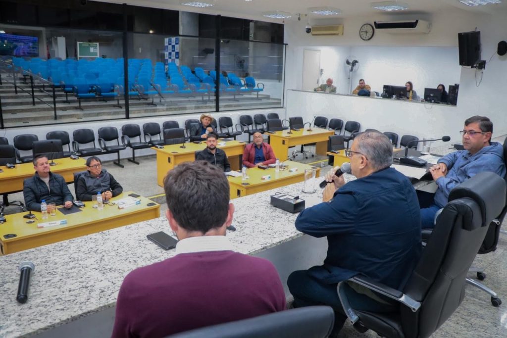 Vereadores no Plenário observam secretário Claudinei Galo, na Mesa Diretiva, falar ao microfone. Ao lado dele, estão o vereador Denis (à direita da foto, de óculos) e o diretor de Trânsito, Rafael Pacheco.