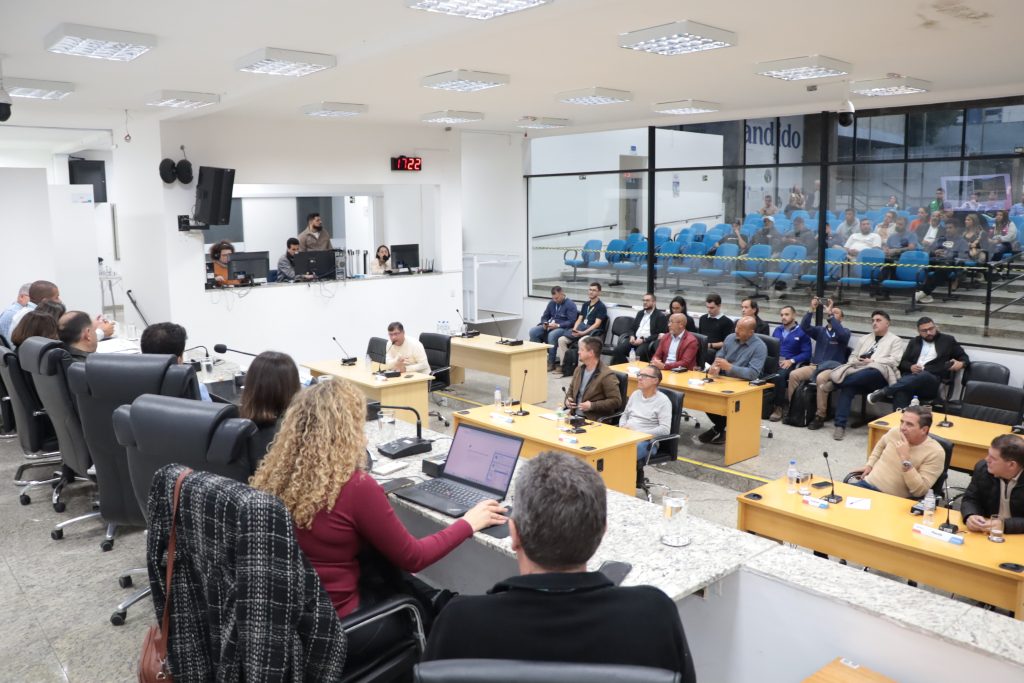em primeiro plano, estão, de costas, na Mesa Diretiva da Câmara, representantes da Sabesp e vereador Marcio Malt. Em segundo plano, de frente para a câmera, estão vereadores e funcionários da Sabesp no Plenário.