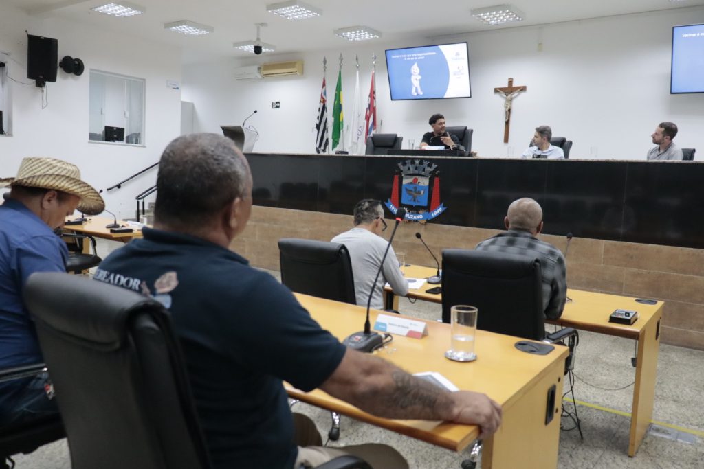 em primeiro plano, vereadores de costas acompanham em suas mesas apresentação da Secretaria de Saúde em audiência realizada em novembro de 2025. Em segundo plano, de frente para a câmera, estão na Mesa Diretiva, da esquerda para a direita, vereador Rogerio Castilho, ex-secretário Diego Ferreira e enfermeiro José Landim.