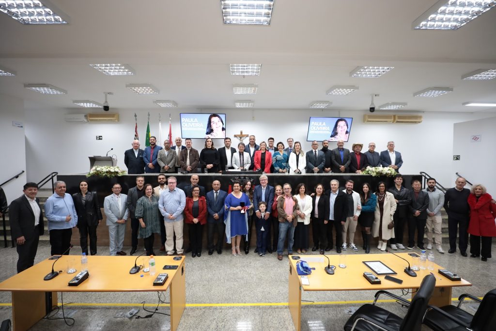 de pé, no Plenário da Câmara, a homenageada Paula Oliveira posa para foto com a Medalha Suzano Brandão rodeada de familiares e amigos. Acima deles, estão na Mesa Diretiva vereadores, orefito e demais autoridades presentes ao evento.