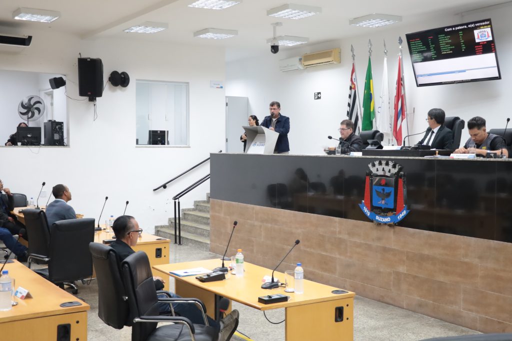 Vereadores sentados atrás de suas mesas no Plenário e vereador Denis falando ao microfone na Tribuna.