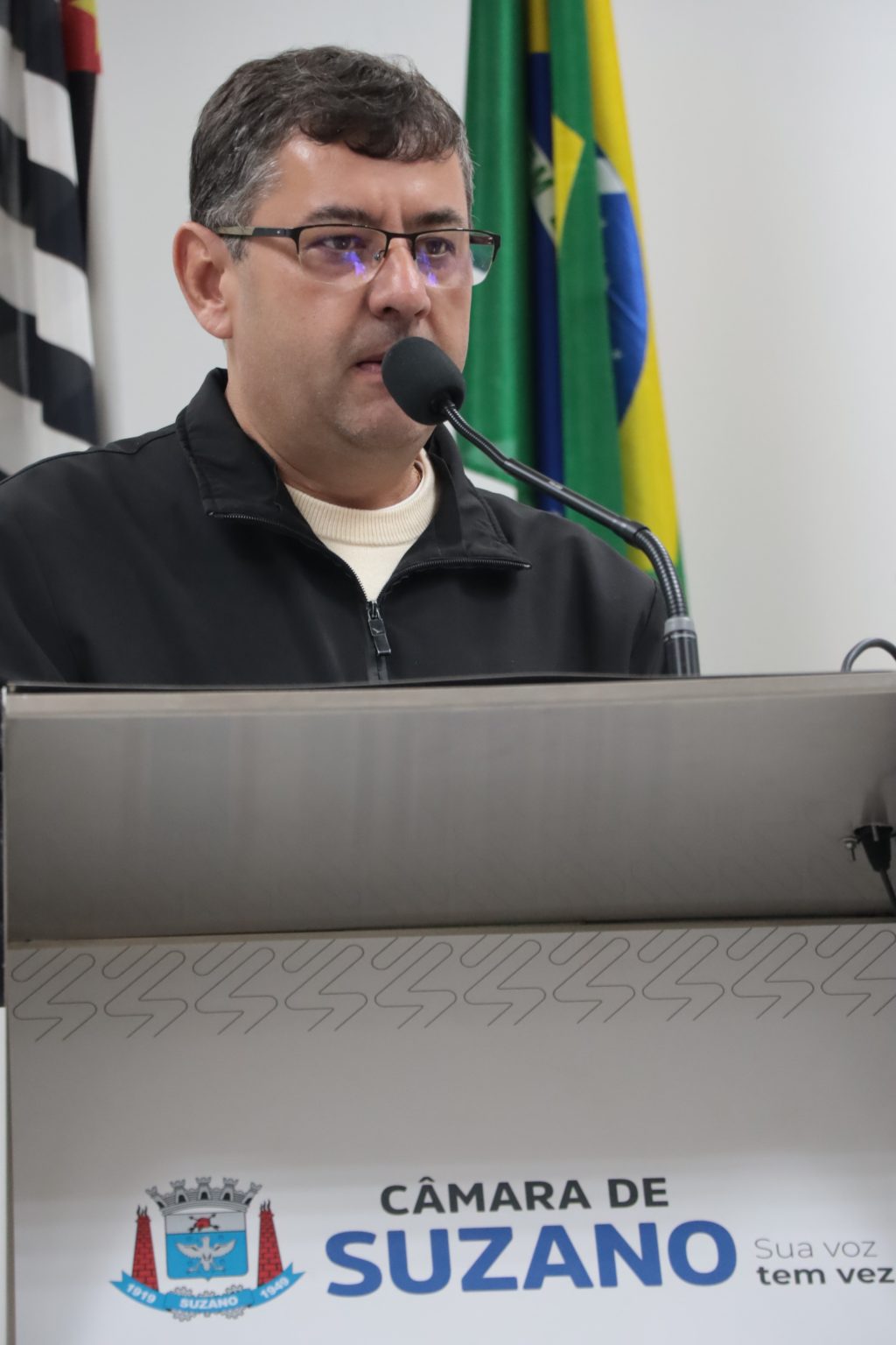 vereador Denis fala ao microfone na Tribuna da Câmara. Ele usa óculos e jaqueta preta.