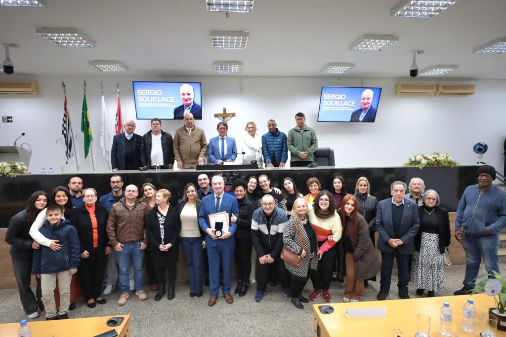 de pé, no Plenário da Câmara, Sérgio Squillace segura a Medalha Antônio Marques Figueira rodeado por familiares e amigos. Acima deles, na Mesa Diretiva, estão vereadores e demais autoridades presentes no evento.