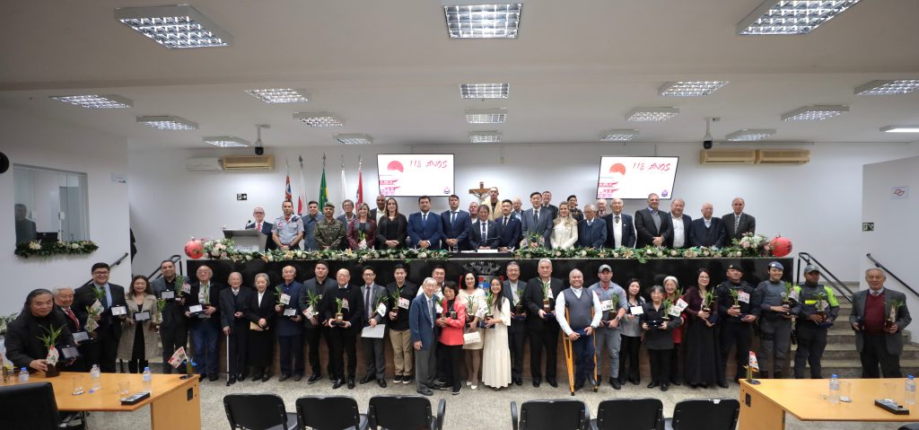 de pé, no Plenário da Câmara, homenageados posam para foto com as placas recebidas. acima deles, na Mesa Diretiva, estão vereadores, prefeito e demais autoridades presentes ao evento.