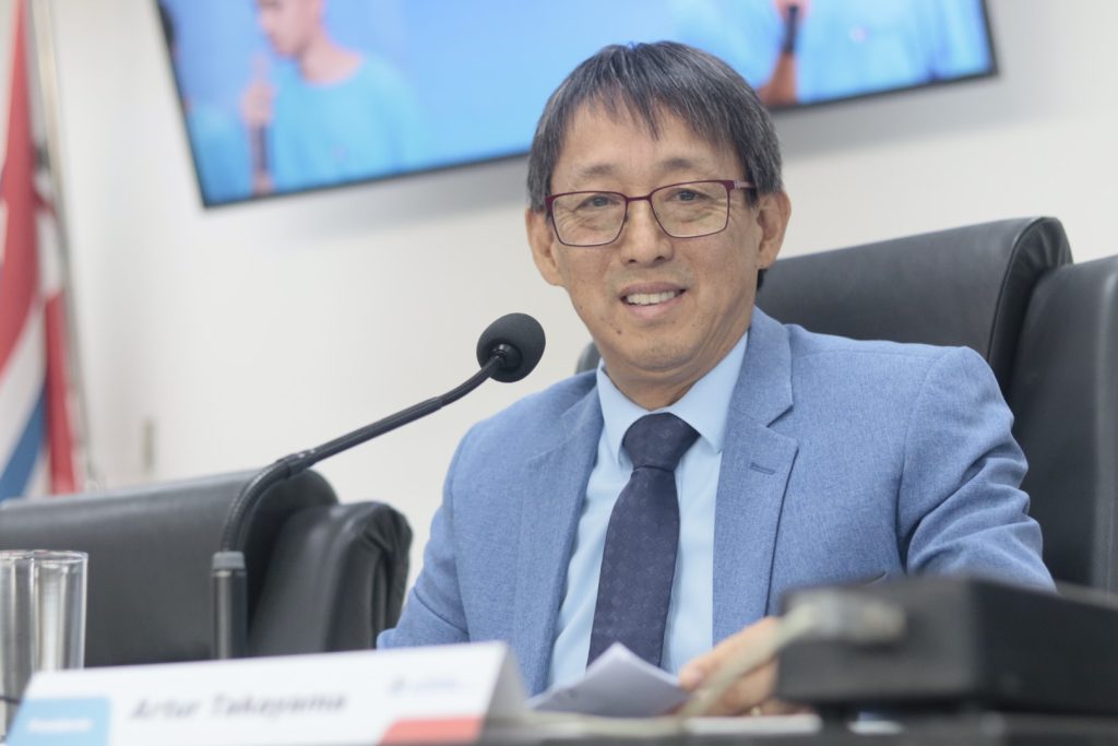 vereador Artur Takayama sorri para foto sentado em sua cadeira na Mesa Diretiva. Ele usa terno e gravata azuis.