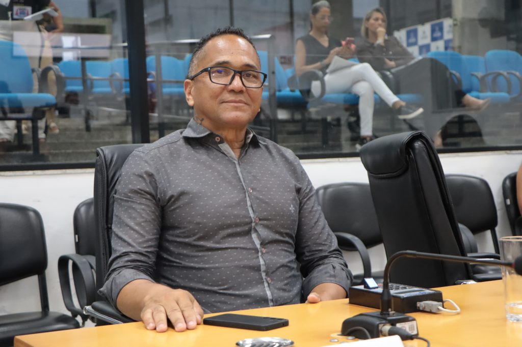 vereador Inhame sentado atrás e sua mesa no Plenário da Câmara. Ele usa óculos e camisa social cinza.