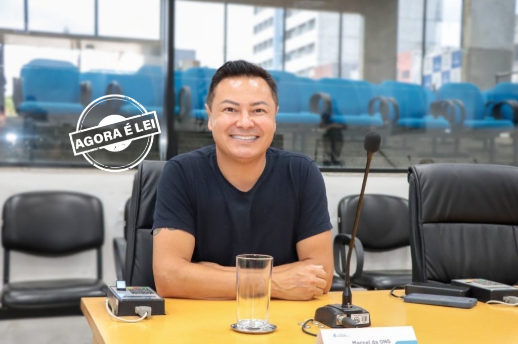  vereador Marcel da ONG sentado atrás de sua mesa no Plenário da Câmara. Ele usa camiseta escura e sorri para a foto.