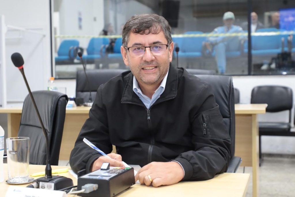 vereador Denis sentado atrás de sua mesa no Plenário da Câmara. Ele usa óculos e jaqueta preta.