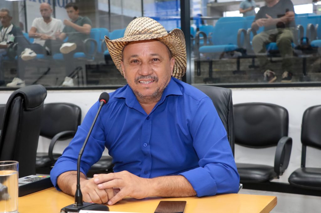 vereador Zé Oliveira sentado atrás de sua mesa no Plenário da Câmara. Ele usa chapéu e camisa social azul.