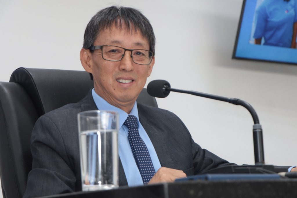 vereador Artur Takayama sentado na Mesa Diretiva do Plenário da Câmara. Ele usa óculos, terno escuro e gravata azul.