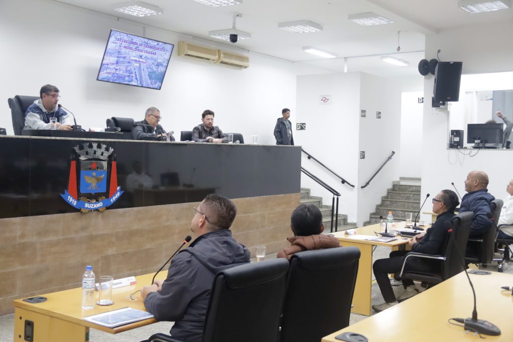 vereadores de costas, sentados atrás de suas mesas no Plenário da Câmara. De frente para eles, na Mesa Diretiva, estão, da esquerda para a direita, o vereador Denis, o secretário Claudinei Galo e o coordenador Rafael Martins Pacheco.
